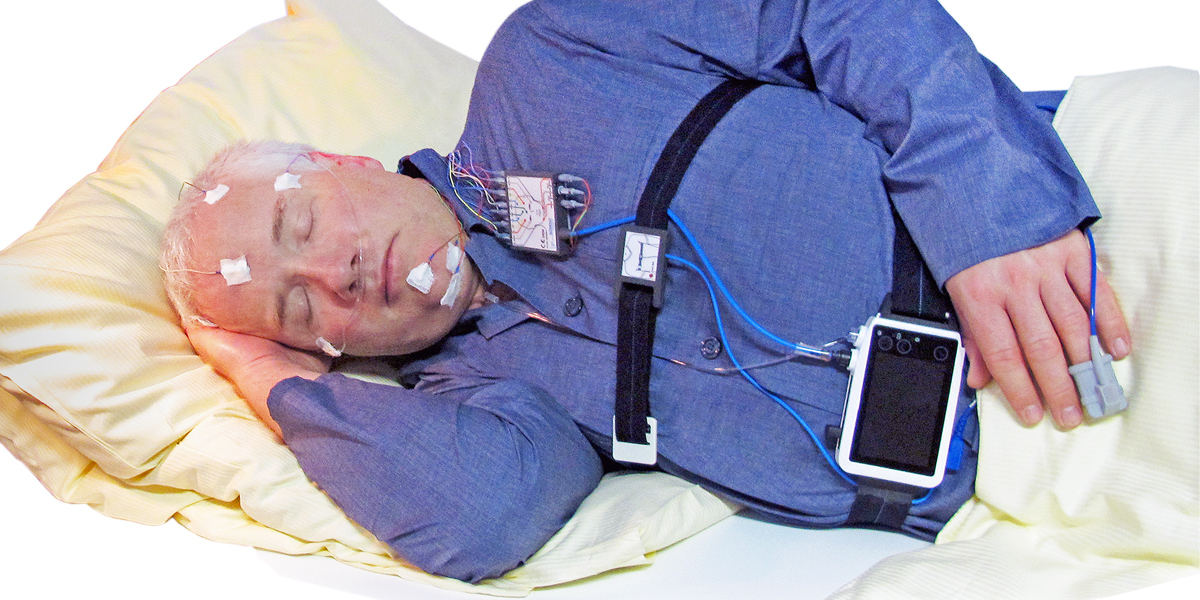 A patient is sleeping wearing a PSG, Polysomnography sleep diagnostic device called SOMNO HD from Somnomedics. The picture shows a built in effort belt and a very small EEG head box. The PSG device is very small and the cables run behind the belt- 